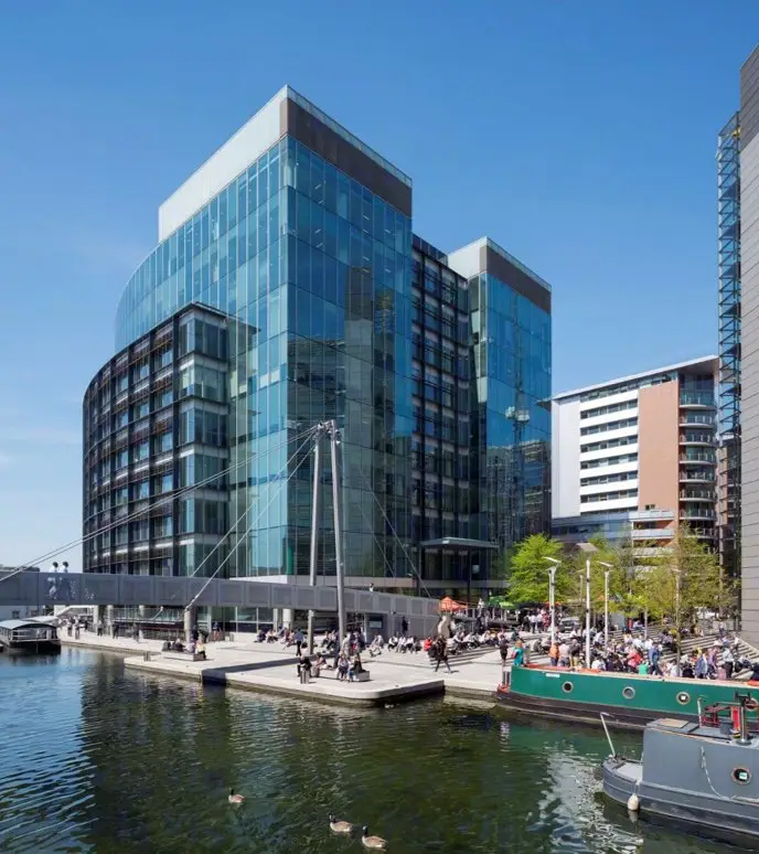 Modern glass office buildings along a waterfront with people near the water.